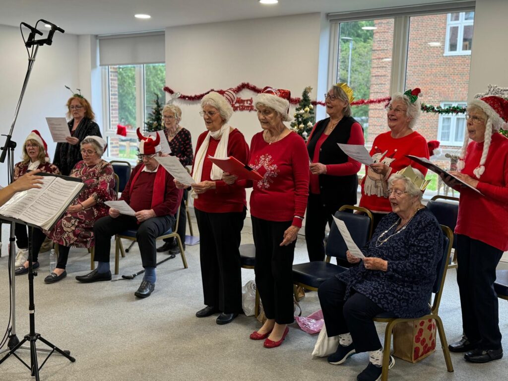 A choir of older people wearing red Christmas jumpers, sing in a room decorated with tinsel. A microphone is visible over their heads, and they are turned towards a music stand to one side